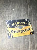 Photo showing the rectangular metal Harley Davidson wall sign with flame design, packaged in plastic sleeve on dark wood surface.