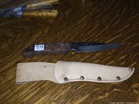 Photo of vintage hunting knife with wooden handle lying next to its sheath on a wooden surface.