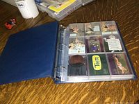 Blue ring binder open, displaying multiple rows of Pepsi collector cards in plastic sleeves on a wooden surface.