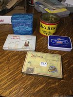 Five vintage tobacco tins arranged on a wooden surface including Edgeworth, Big Ben, Sweet Caporal, Rothmans, and Sportsman brands with visible wear and aging.