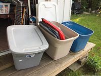 Three plastic storage totes in gray, beige, and blue, with the beige one containing several spare lids visible on top.