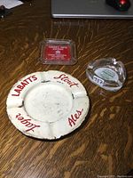 Lot displaying three ashtrays: Labatt's enamel metal ashtray with visible wear, a square glass ashtray commemorating Canada's 100th birthday, and a round clear glass ashtray labeled Lulus.