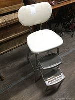 Front view of the vintage kitchen step stool showing the vinyl padded seat and backrest, chrome frame, and two metal steps with signs of wear and rust.