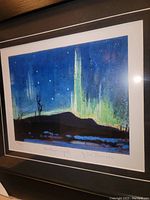 Close-up of framed art print showing night landscape with Northern Lights aurora in green and blue, stars in dark sky, and blue river below. Signed and titled below the print.