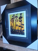 Full view of the framed fine art print showing tall pine trees silhouetted against a sunset backdrop in a black frame with corner protectors.