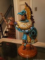 Full view of the Medicine Man sculpture showing the standing pose, intricate feather cape, colored stones, and wooden base.