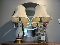 Pair of white ceramic table lamps with gold and green floral patterns, brass bases, and cream scalloped shades, placed in front of a circular mirror.