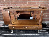 Front view of walnut bar cart showing top shelf, central tray, lower shelf, drawers and casters