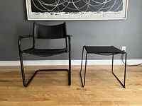 Front view of black leather cantilever chair with black spaghetti style stool beside it on wood floor under wall art.