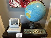 Photo of all three items on a black wooden surface: vintage Paymaster cheque writer, Lightning adding machine, and illuminated globe.