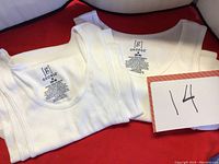 Two folded white undershirts laid out side by side on a red background showing the front neckline area and the undershirt shape.