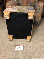 Bottom view of the ottoman showing wooden block feet with white knobs and underside fabric in black, placed next to matching chair.