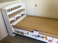 Twin bed frame with white and natural wood headboard with built-in shelves and three pull-out drawers under the mattress platform with colorful letter stickers on front of drawers spelling 'Wish It' and 'Dream It'.