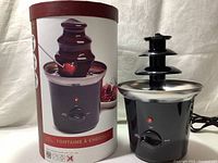 Image showing the Decora chocolate fountain next to its cylindrical packaging with product picture and label.