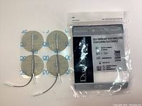 Four round 2 inch electrodes with lead wires laid out next to sealed original package of Dura-Stick self-adhesive stimulating electrodes.