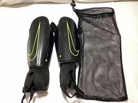 Pair of black Nike youth shin guards with neon yellow Swoosh logos and attached black elastic socks with toe loops, lying flat next to black mesh pouch.