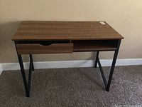 Front view of woodgrain desk with black metal legs, showing one drawer and open cubby below top surface.