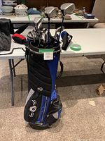 Full view of the black and blue golf bag with clubs inside, showing 'John Daly' embroidered on the bag front pocket.