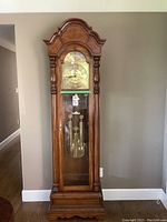 Front view of oak grandfather clock showing arched top, glass door, clock face with Roman numerals, and brass pendulum and weights inside.
