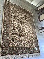 Full view of rectangular wool rug with cream field and floral designs bordered by green patterned edge and fringe tassels at the ends.