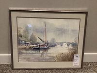 Framed watercolour painting depicting a calm riverside scene with boats, a stone arch bridge, and trees in the background.