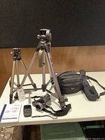 Full view of two aluminum tripods, camera bag, and Panasonic video camera on table with tag.