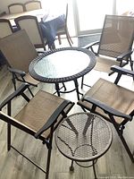 Overall group shot of patio table with glass top, four folding chairs with beige woven fabric, and small round metal mesh end table.