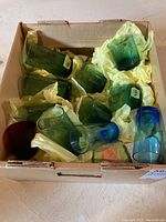Box of assorted coloured glass tumblers in green, blue, and red wrapped with yellow tissue paper inside cardboard box.