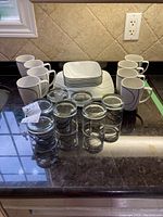 Photo shows 8 white mugs with black curved line designs alongside 6 clear glasses with black swirl designs, and 6 square bowls with black lines, arranged on the counter.