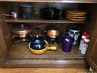 Shelf with various fondue pots including Le Creuset yellow and blue fondue pot, copper and brass pots, stacked yellow ceramic plates, metal burner stands, and butane fuel cans