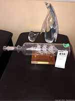 Clear glass art items including a penguin, a bird, and a ship in a bottle on a wood base, photographed on a black table against a beige wall.