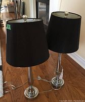 Pair of chrome base table lamps with black fabric tapered drum shades, set on hardwood floor.