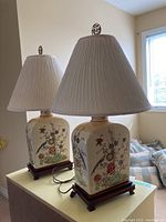 Pair of matching porcelain table lamps with wooden bases and cream fabric shades shown on a table by a window.