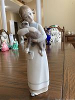 Full figure shot of Lladro figurine showing girl holding lamb on wooden table, other figurines and home decor in background