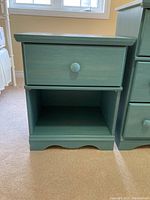 Front view of a green painted wood nightstand with a drawer and open shelf below, sitting on carpet.