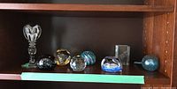 Shelf showing 8 decorative paperweights including glass bird, gold and blue patterned pieces