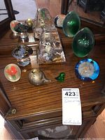Top view of all paperweights arranged on wooden surface showing various shapes and colors including mushroom, flower, green egg shapes, dandelion, and triangular acrylic paperweights.