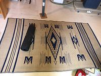 Full view of large outdoor patio carpet laid flat with cone shaped legs of furniture and wooden shelving visible in background, carpet has central diamond and symbolic pattern in dark blue on tan base with decorative edging.