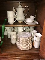 Wide shot of cabinet shelves showing coffee pot, stacked mugs, plates, saucers and serving tray