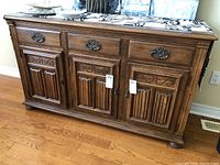 Front view of brown wooden sideboard with three drawers and three cabinet doors, adorned with ornate metal handles and decorative carved panels