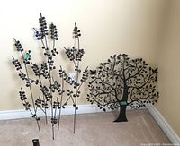 Two black steel wall art pieces resting against a wall on carpeted floor, showing tree and branch motifs with leaf clusters.