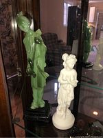 Green translucent Asian figure and white classical female figure displayed on glass shelf with mirror reflection.