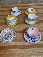 All six teacups with matching saucers arranged on wooden table showing various patterns and colors.