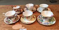 Six teacups and saucers arranged on a wooden surface with varied floral and fruit designs