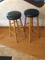 Set of two wooden bar stools with black faux leather seats shown from side view on wooden floor.