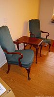 Two green upholstered chairs with dark wood arms and legs surrounding a dark wood rectangular accent table on a wooden floor. The furniture has visible scratches and requires cleaning.