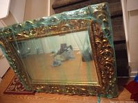 Large ornate gold framed mirror wrapped in protective plastic with visible minor frame damage