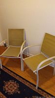 Two light brown canvas patio chairs with light metal frames and armrests on wooden floor near wall and carpet.