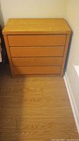 Photo of light brown 4-drawer chest with visible scratches on drawers and top, positioned against a wall on wooden floor.