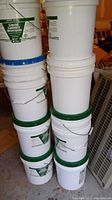 Photo showing stacked white plastic 5-gallon drums with various colored lids including green, blue, and orange, each with pouring spouts